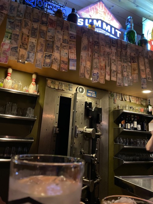 A bar interior featuring a wall of money, shelves of drinks, and a large safe door in the background.