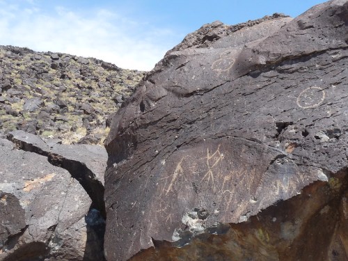 Petroglyphs etched into a large rock, surrounded by rocky terrain and a clear blue sky.