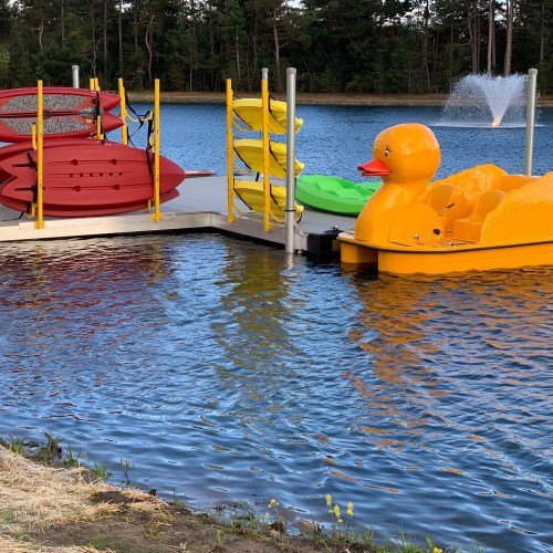 Colorful paddle boats and a large rubber duck float at a dock by a serene lake, surrounded by trees.