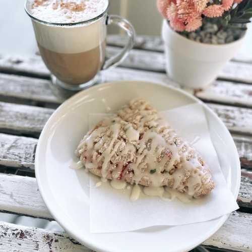 A plate with a glazed pastry and a latte in a glass mug, alongside a small potted plant on a rustic table.