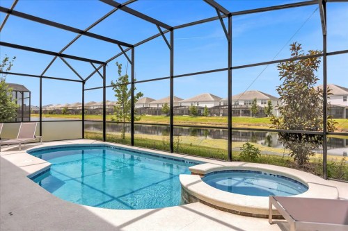 A serene pool area with a hot tub, enclosed by a screen, overlooking a calm lake and houses in the background.