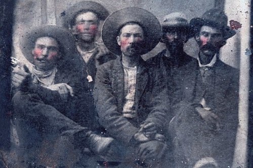 A vintage photo of five men in hats, with painted red cheeks, sitting together in a rustic setting.