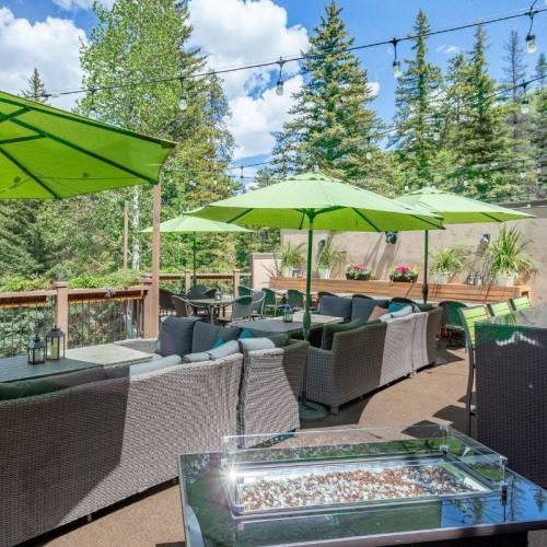 A cozy outdoor lounge area with green umbrellas, comfortable seating, and a fire pit, surrounded by trees and blue skies.