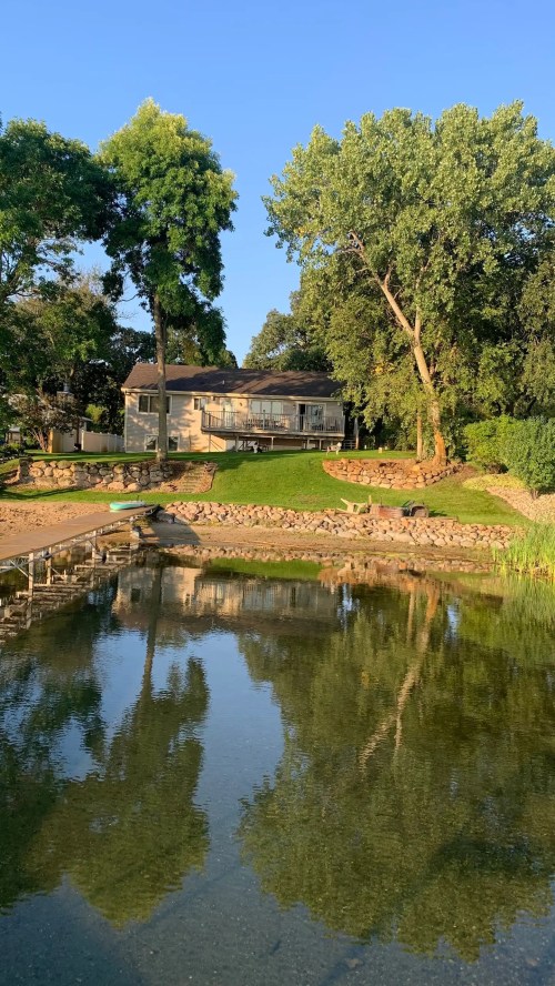 A serene lakeside view featuring a house, trees, and reflections on the water under a clear blue sky.