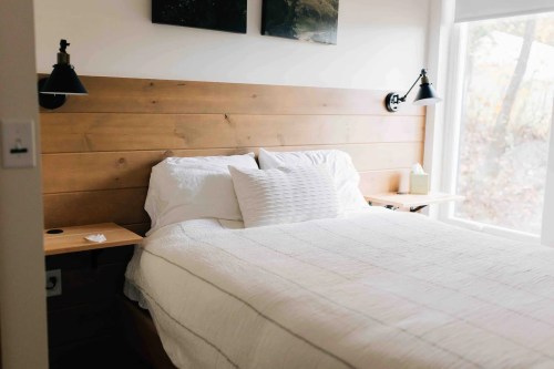 Cozy bedroom with a wooden headboard, white bedding, and bedside lamps, featuring large windows with natural light.