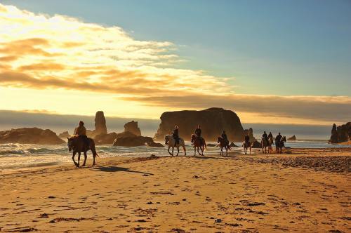 A group of people riding horses along a sandy beach at sunset, with rocky formations in the background.
