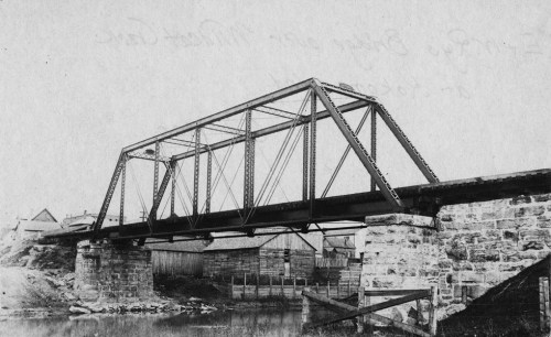 Black and white photo of a metal truss bridge over a river, with stone supports and nearby buildings.