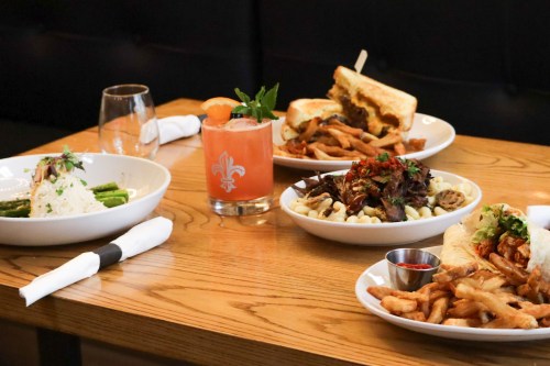 A table with four dishes: rice with asparagus, fries with meat, a sandwich, and a cocktail, all on a wooden surface.