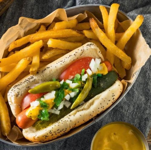 A hot dog with toppings in a bun, served with golden fries and a small cup of mustard.