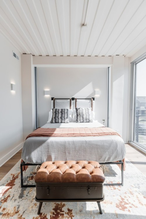Modern bedroom with a double bed, decorative pillows, a tufted bench, and large windows letting in natural light.