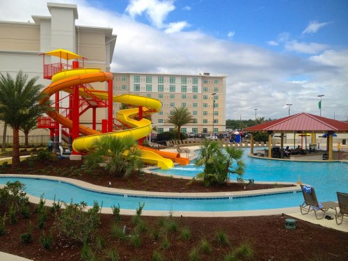 A colorful water slide next to a pool, surrounded by palm trees and a hotel in the background under a cloudy sky.