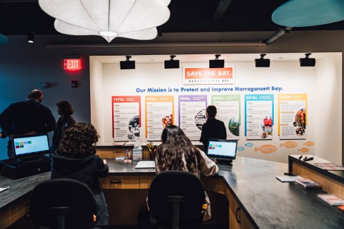 A reception area with people at computers, featuring a wall display about protecting Narragansett Bay.