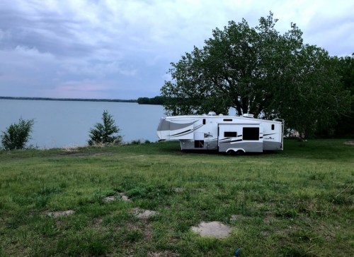 A parked RV by a lake, surrounded by green grass and trees under a cloudy sky.