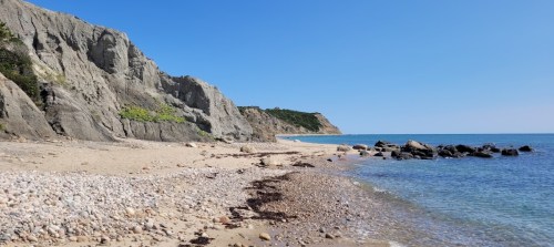 A serene beach scene with rocky cliffs, clear blue water, and a sandy shore under a bright blue sky.