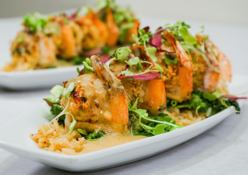 A plate of shrimp topped with sauce, served on a bed of rice and garnished with greens and microgreens.