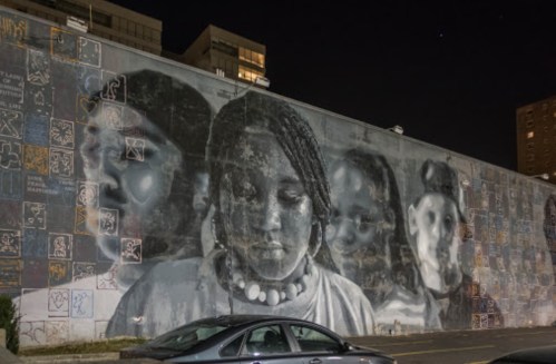 A large mural at night featuring portraits of four young people, with colorful graffiti surrounding the images.