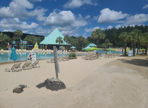 A sandy beach area with lounge chairs, umbrellas, and a pool surrounded by palm trees and blue skies.