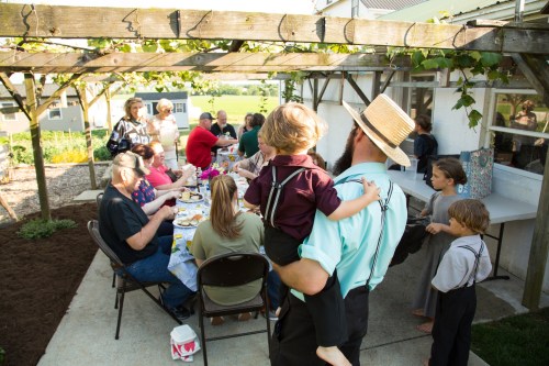 A gathering of people enjoying a meal outdoors under a pergola, with children and adults seated at a long table.