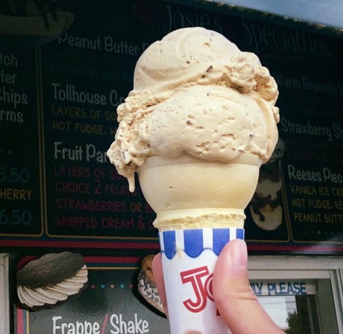A hand holding a large cone of creamy peanut butter ice cream in front of an ice cream shop menu.