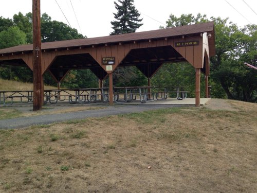 A wooden pavilion with picnic tables, set in a grassy area surrounded by trees.