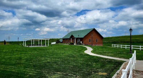 A wooden house with a green roof surrounded by grassy fields and a pathway, under a cloudy sky.