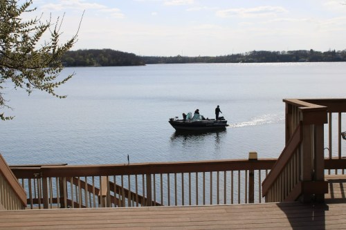 A small boat with two people glides across a calm lake, framed by wooden steps and trees in the foreground.