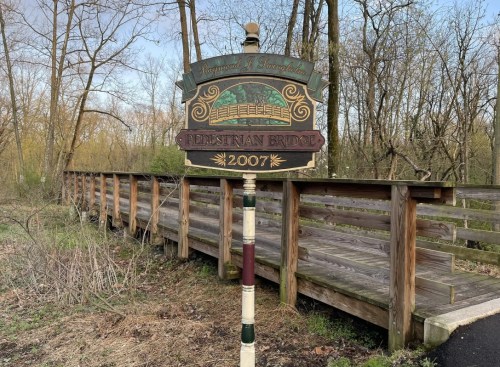 Sign for a pedestrian bridge, surrounded by trees, with a wooden walkway visible in the background.