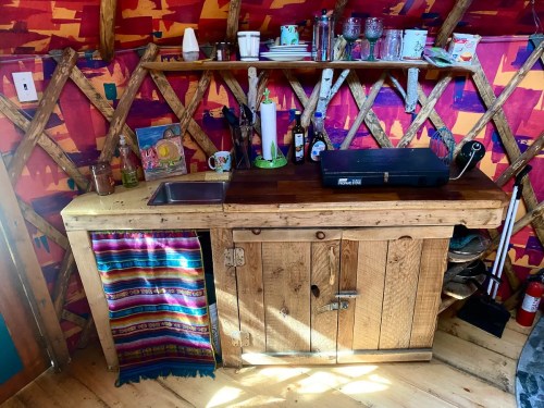 A rustic wooden kitchen counter with a sink, shelf, and colorful wall decor in a cozy yurt setting.