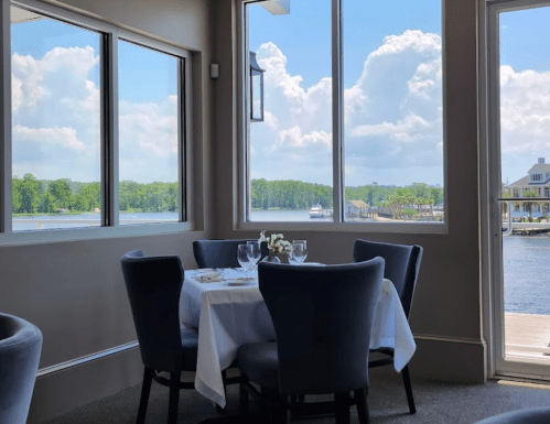 A dining table set for two by large windows, overlooking a scenic waterfront with trees and clouds in the background.