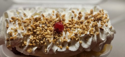 A dessert topped with whipped cream, crushed nuts, and a cherry on a banana split in a takeout container.