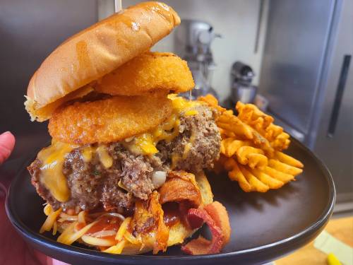 A towering burger with cheese, onion rings, and bacon, served with waffle fries on a black plate.