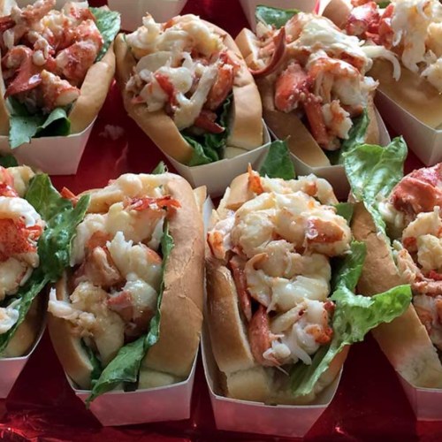 A close-up of lobster rolls filled with lobster meat and lettuce, arranged in white containers on a red surface.