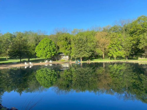 A serene pond reflecting lush green trees and a clear blue sky, with chairs by the water's edge.