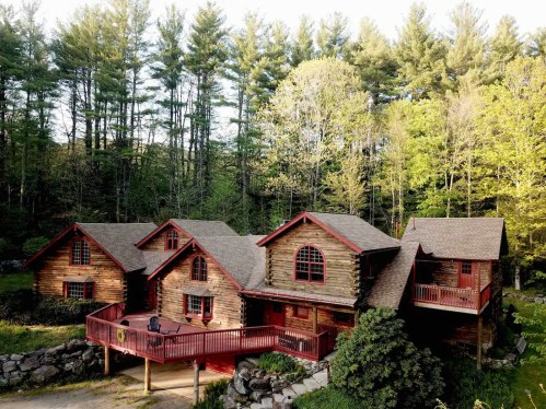 A rustic log cabin surrounded by lush green trees, featuring a large deck and multiple gabled roofs.
