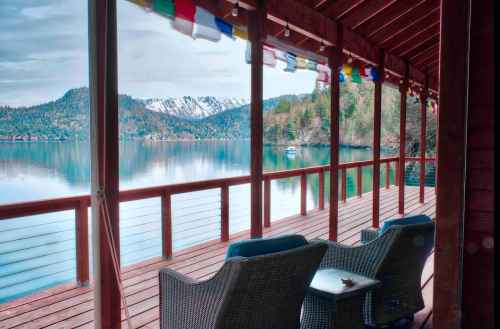 A serene lakeside view from a wooden deck, featuring chairs and colorful prayer flags, with mountains in the background.