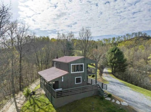 Aerial view of a rustic house surrounded by trees and a gravel driveway under a cloudy sky.