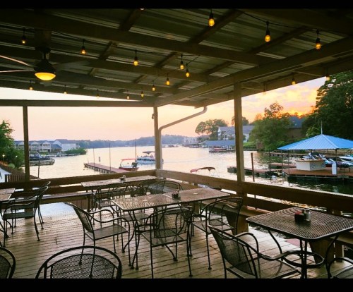 A lakeside restaurant patio with empty tables, string lights, and a view of boats on the water at sunset.