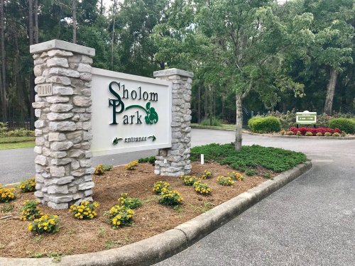 Sign at the entrance of Sholom Park, surrounded by greenery and colorful flowers.