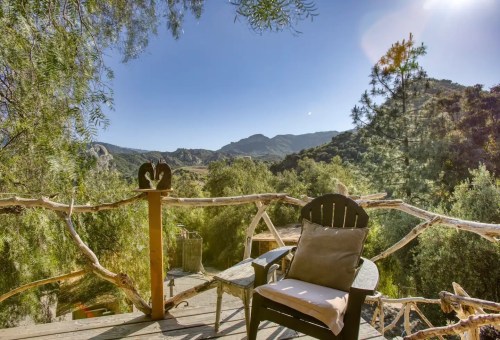 A rustic chair on a wooden deck overlooks a scenic mountain landscape under a clear blue sky.