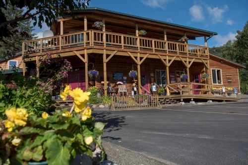 A wooden building with a large porch, surrounded by flowers and greenery, under a blue sky with scattered clouds.
