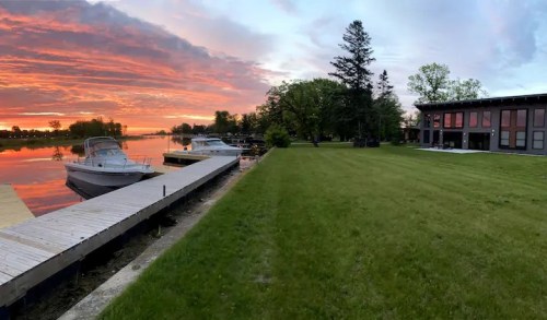 A serene lakeside view at sunset, featuring boats, a wooden dock, lush green grass, and a modern building.