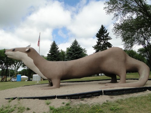 A large, whimsical sculpture of an otter lying on a gravel surface, surrounded by trees and a cloudy sky.