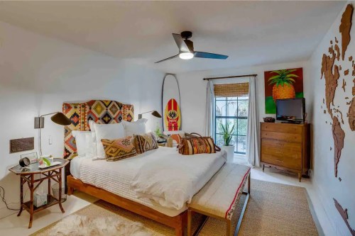 Cozy bedroom with a patterned headboard, surfboard decor, and a window with natural light. Earthy tones and plants.