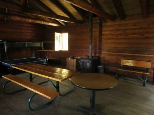 Interior of a rustic cabin featuring wooden walls, a table, benches, a bunk bed, and a wood stove.