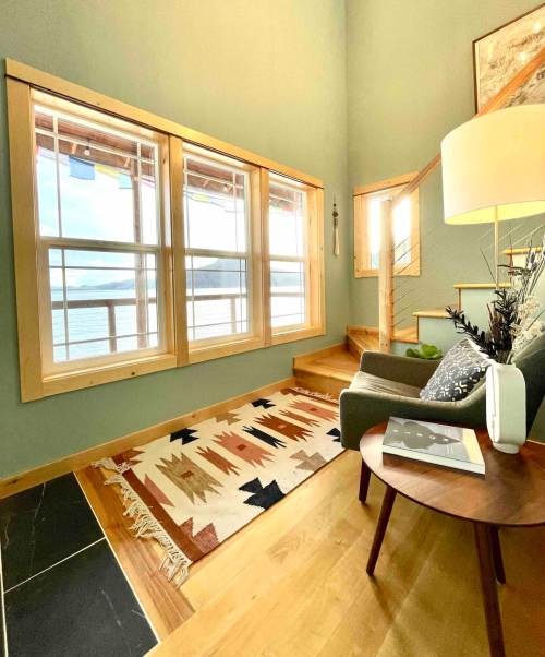 Cozy living space with large windows, a green sofa, wooden stairs, and a decorative rug, overlooking a scenic view.