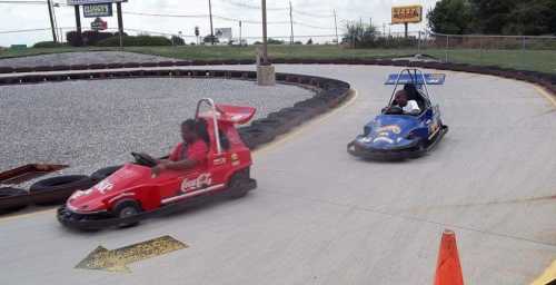 Two children race go-karts on a track, one in a red car and the other in a blue car, with a gravel background.