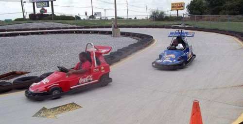Two children race go-karts on a track, one in a red car and the other in a blue car, with a gravel background.