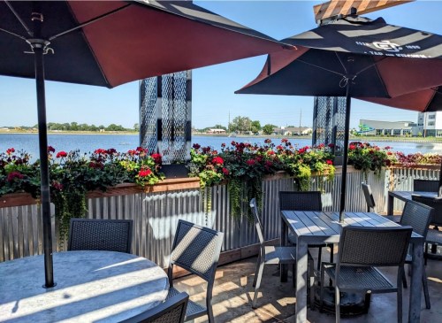 Outdoor dining area with tables, chairs, and umbrellas, overlooking a scenic waterfront with blooming flowers.