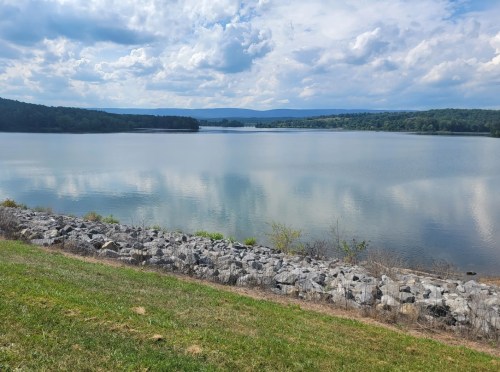 A serene lake surrounded by greenery and rocky shoreline, with clouds reflecting on the water's surface.