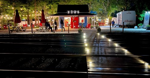 A cozy outdoor scene at night with a red building, picnic tables, and illuminated train tracks.